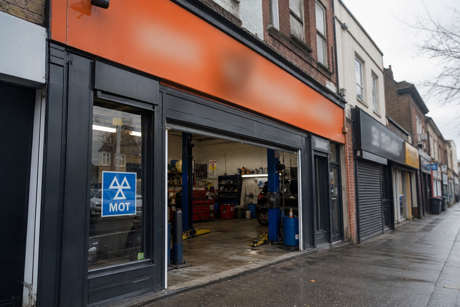 A UK garage fascia in orange, MOT sign in the window, bay door open onto a wet pavement.