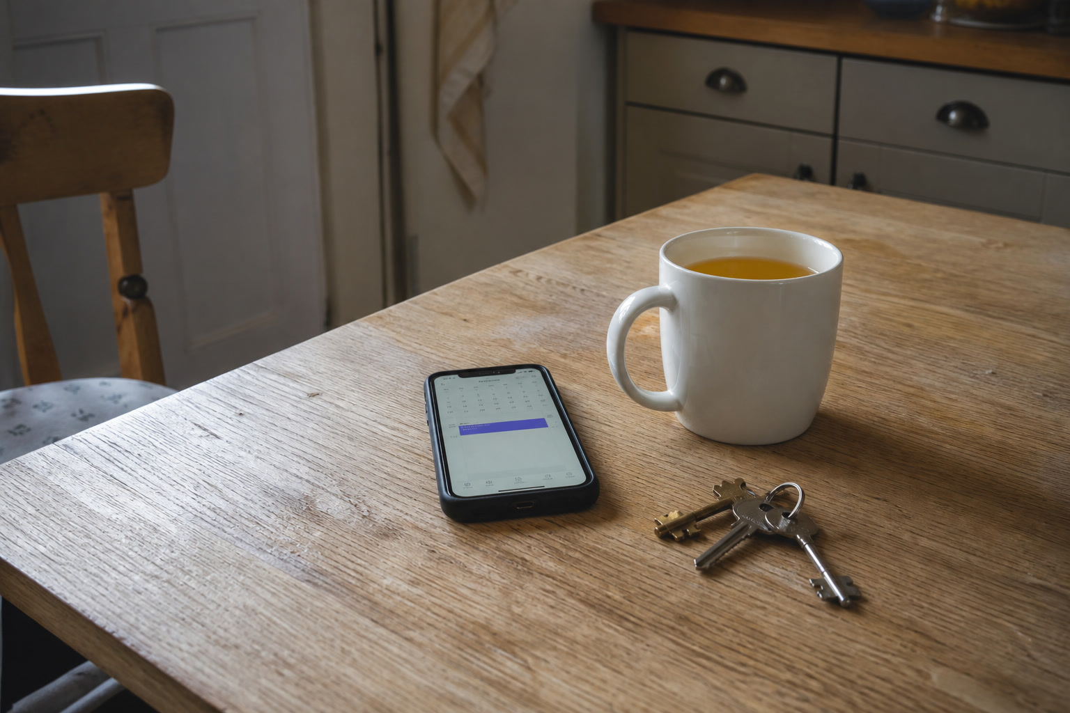 A kitchen table with a phone showing a booking, a mug of tea and car keys.